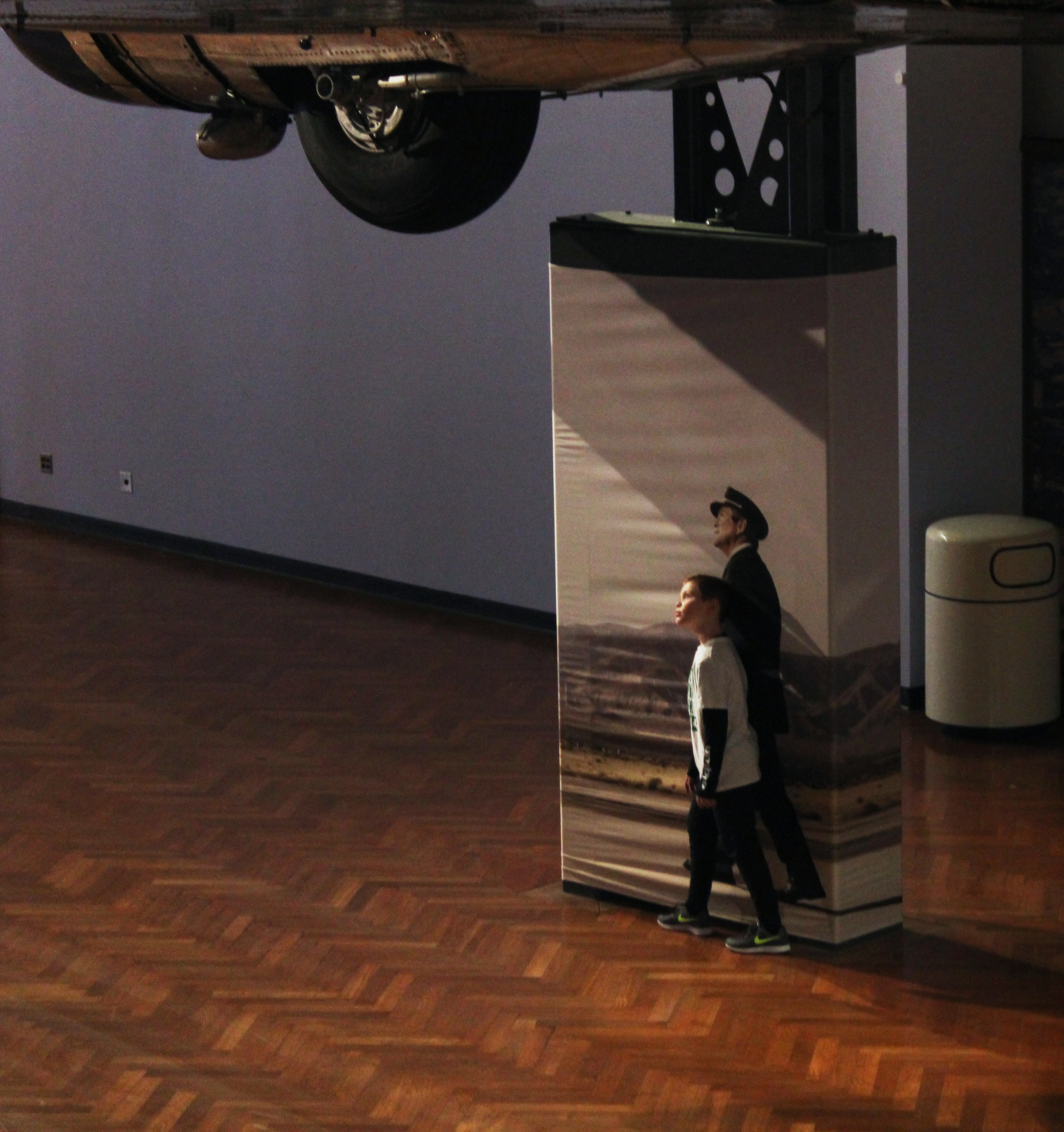 Photo of boy standing next to the image of a pilot as if he is part of the image underneath an airplane hanging at The Henry Ford museum in Dearborn, Michigan.