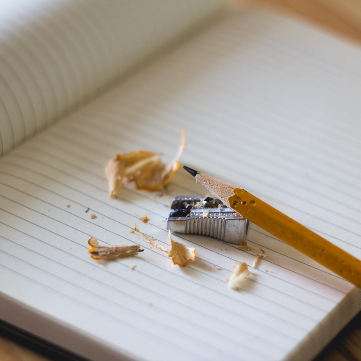 Photo of open notebook with blank lined pages sitting on a desk. On the page is a pencil and pencil shavings. Photo by Angelina Litvin from StockSnap.io