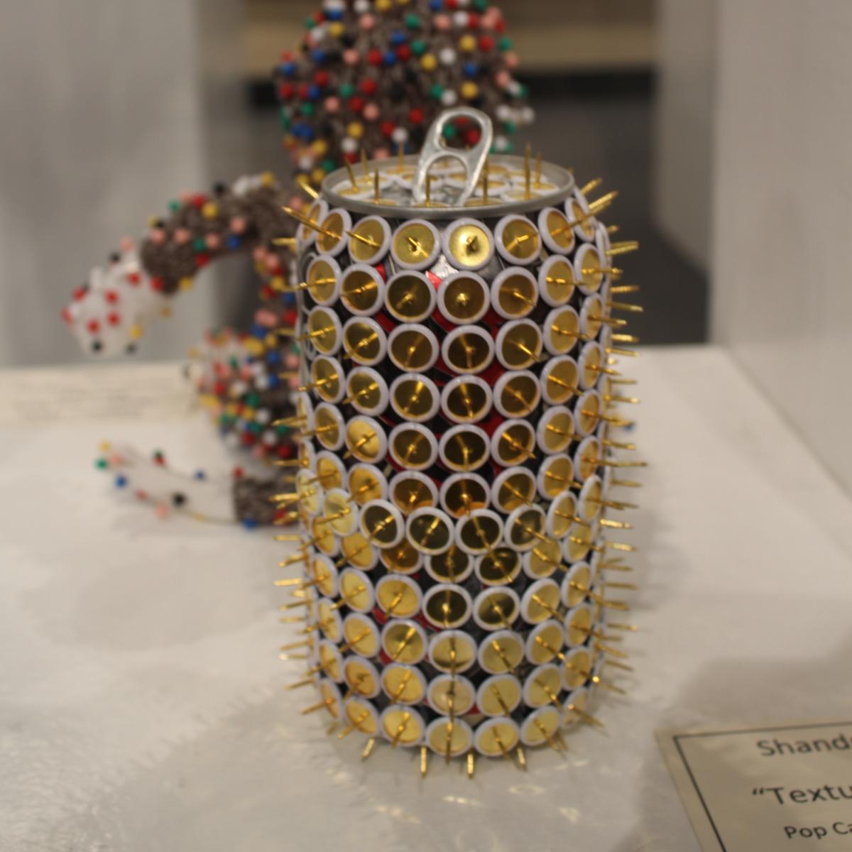 “Texture” by Shandra Dicks - pop can covered with golden thumb tacks sharp end pointed outward.